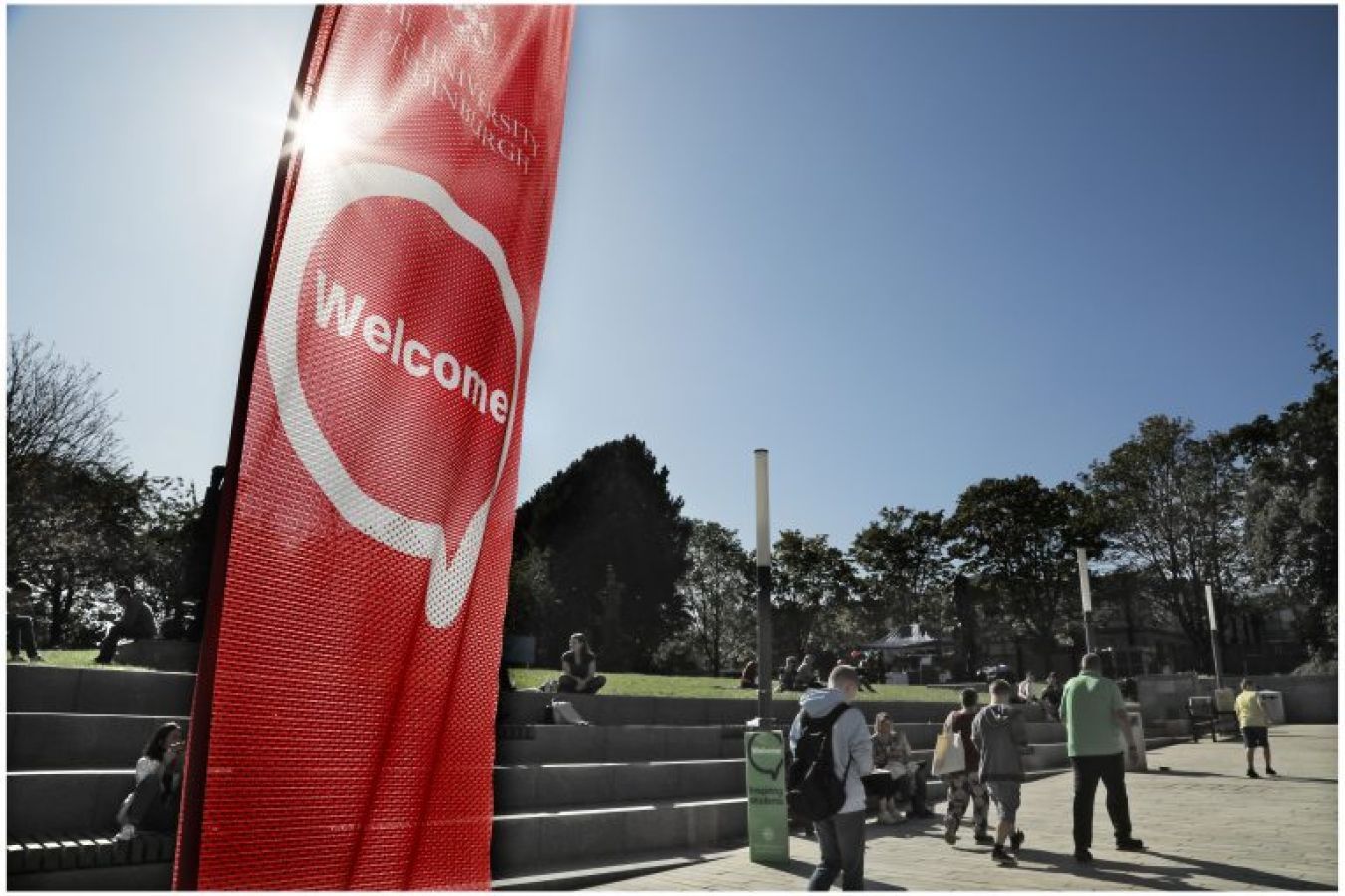 People walking past a red ‘Welcome’ banner in sunshine.