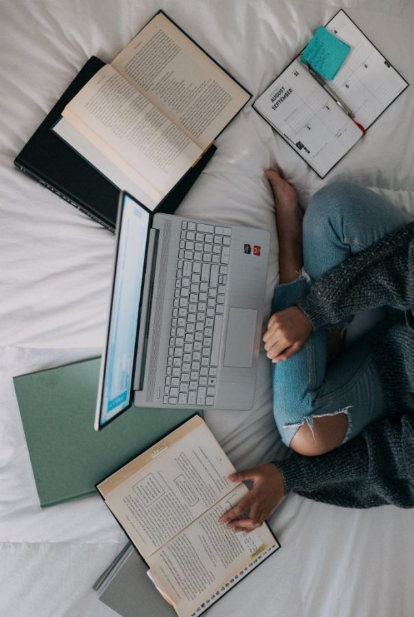 Aerial view of female sitting on a bed surrounded by a diary, books and a laptop