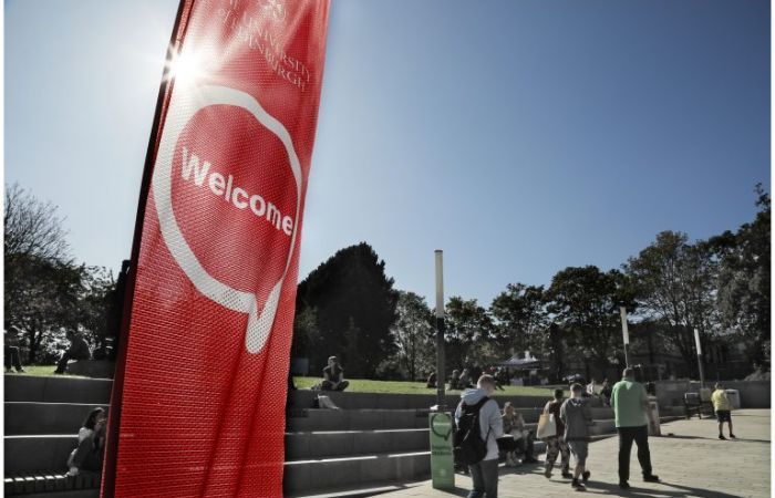 People walking past a red ‘Welcome’ banner in sunshine.